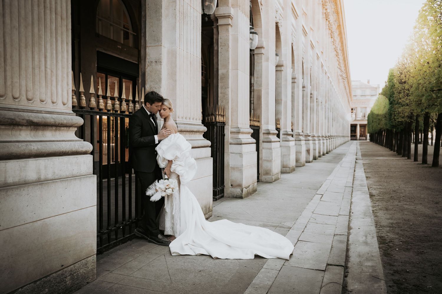 Bride and groom share a romantic moment by ornate stone columns on a sunlit city street.