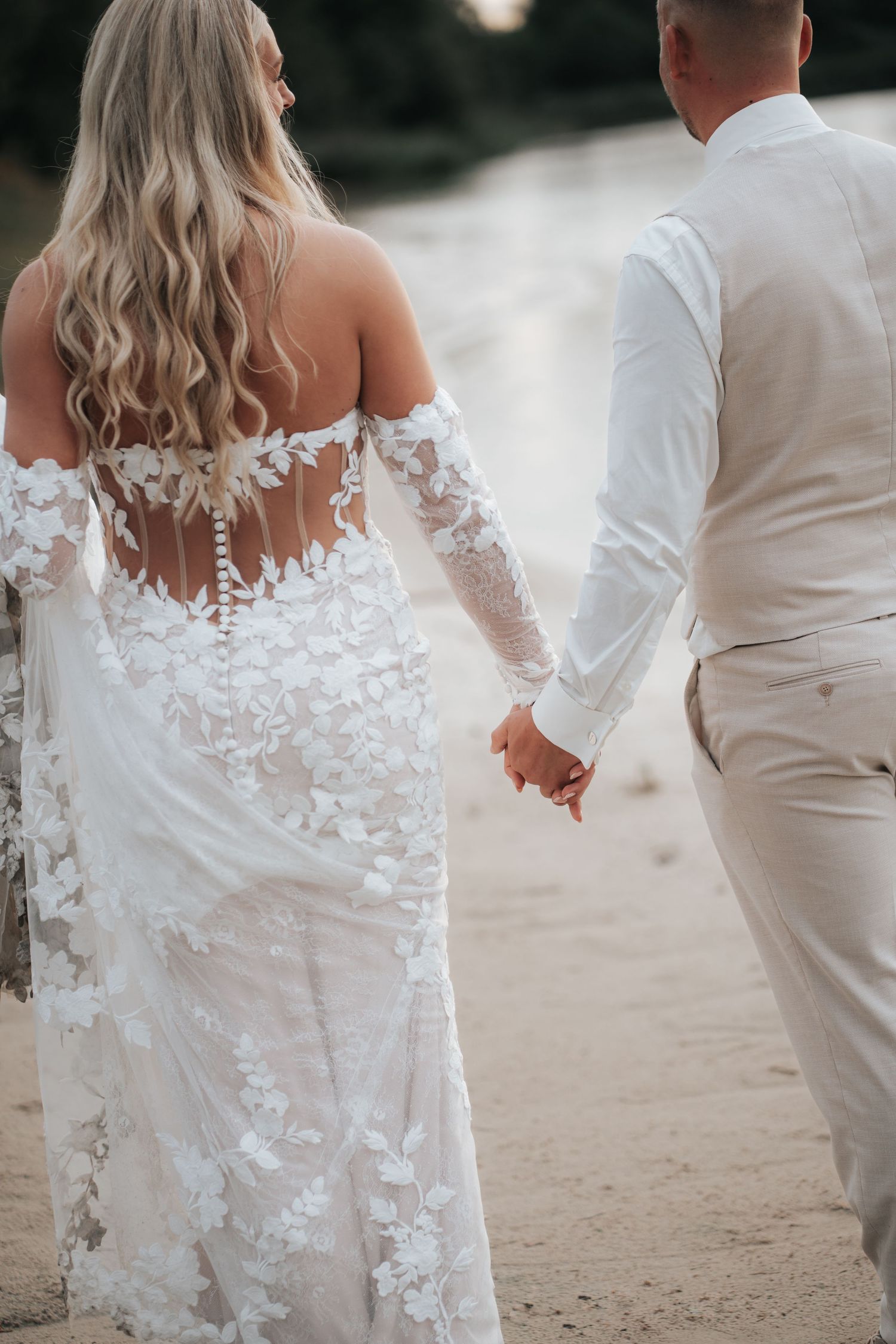 Bride and groom holding hands walking away, bride in stunning backless floral lace wedding gown.