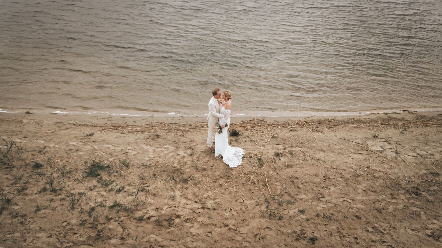 Aerial view of bride and groom embracing romantically on a sandy beach by the water.