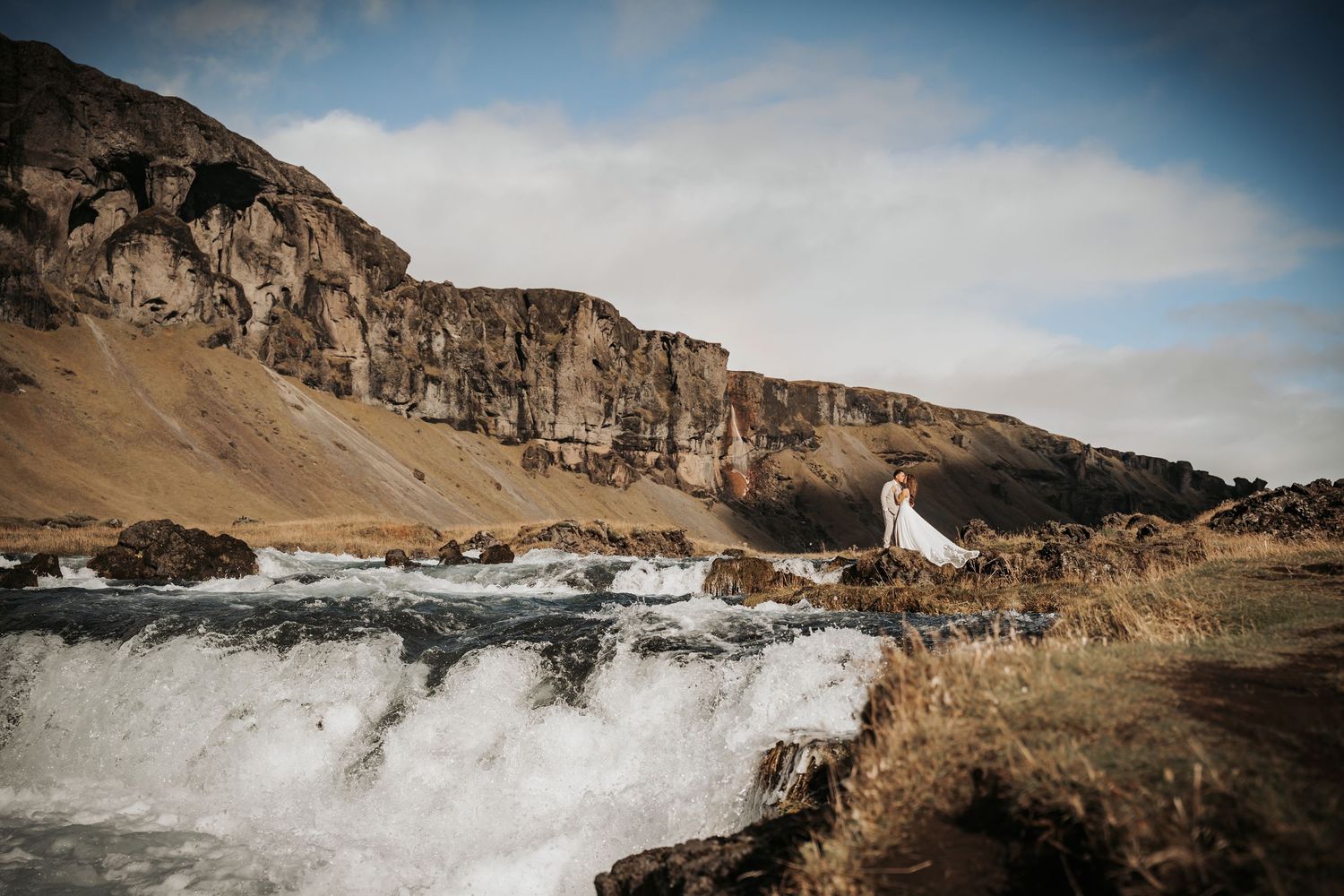 Bride and groom standing near a rushing waterfall with rocky cliffs in the Icelandic highlands.