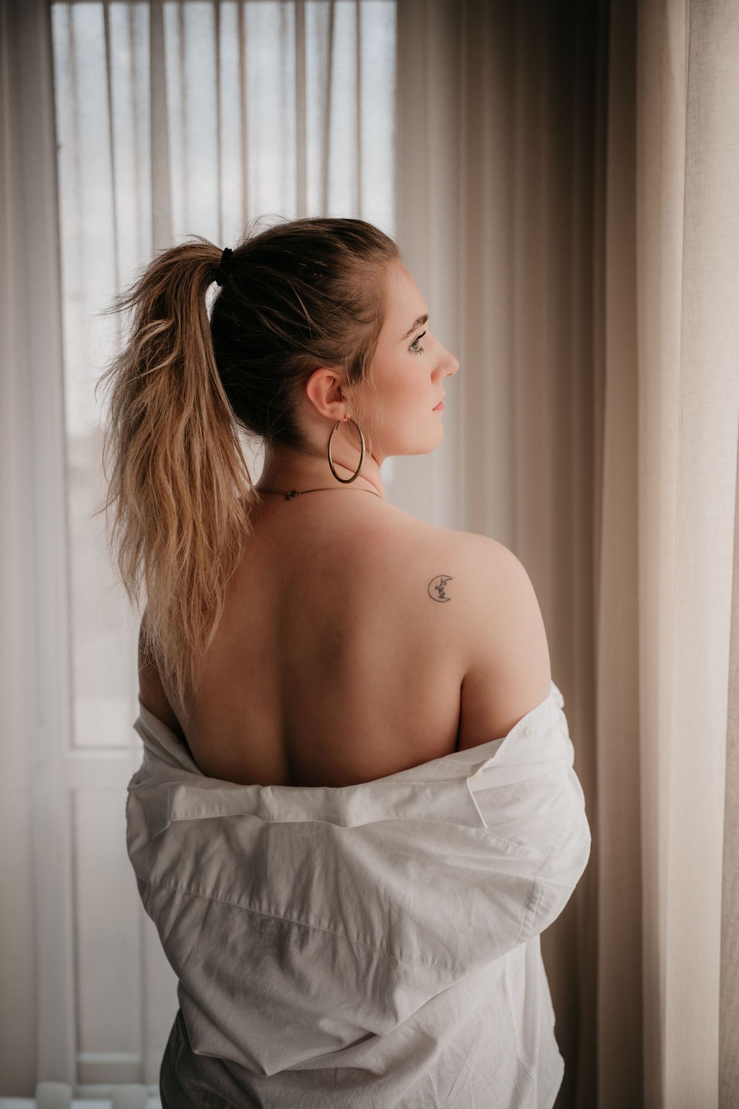 Woman with ponytail and hoop earrings wearing off-shoulder white shirt, showing small moon tattoo on back.