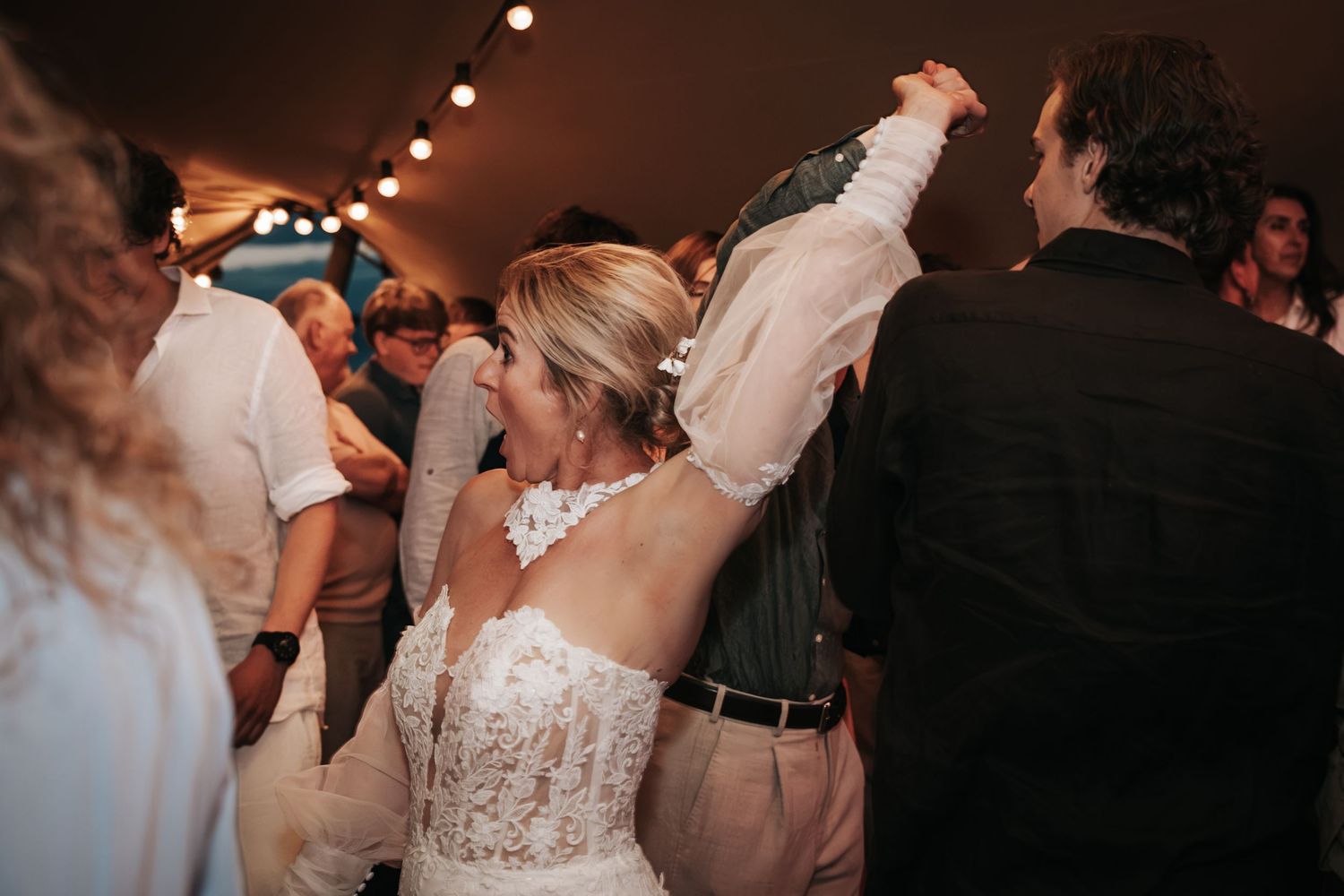 Bride in lace wedding dress dancing with groom under string lights at a wedding reception.