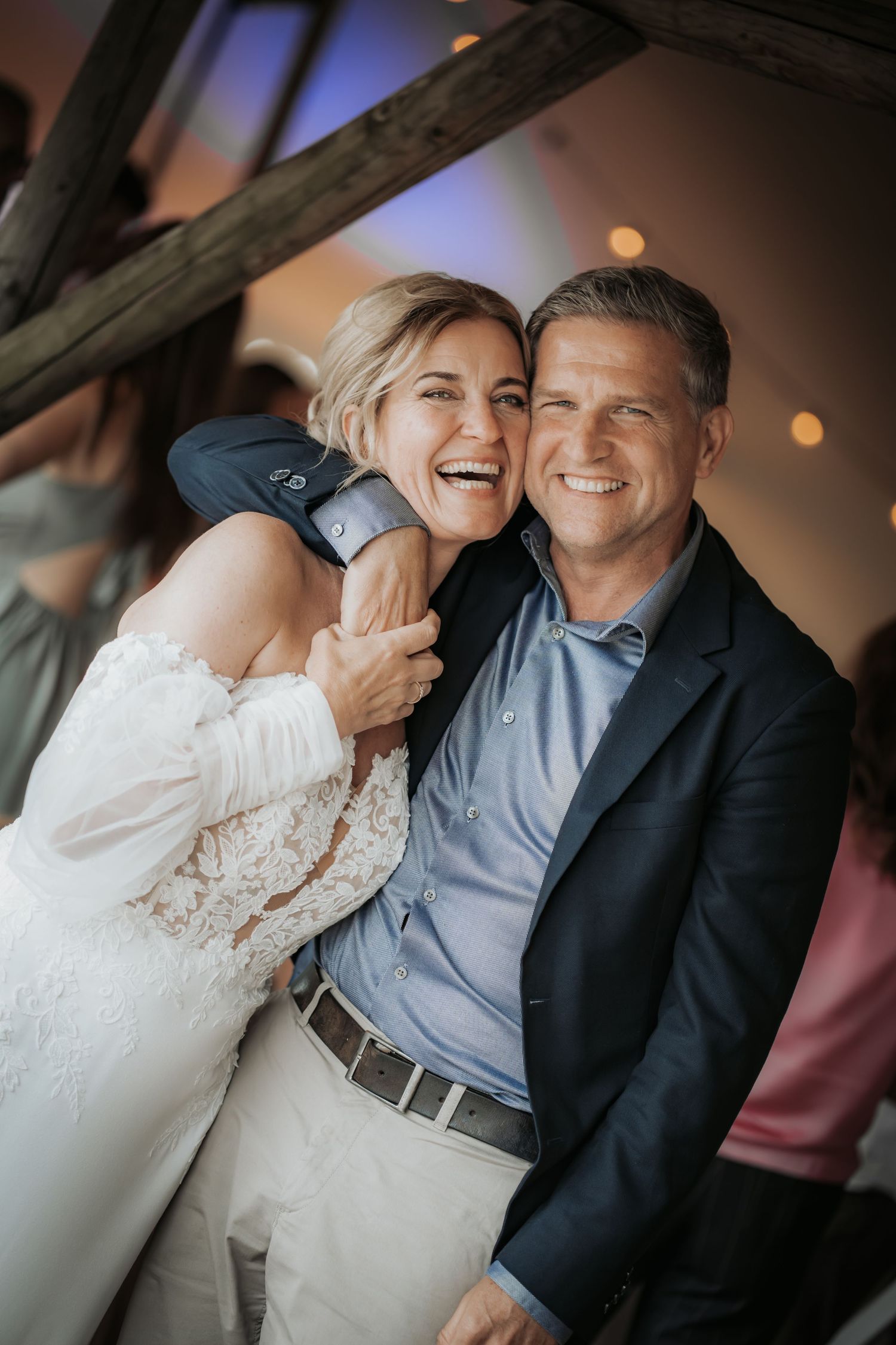Laughing bride in lace dress embraces smiling man in navy blazer at a warmly lit wedding reception.