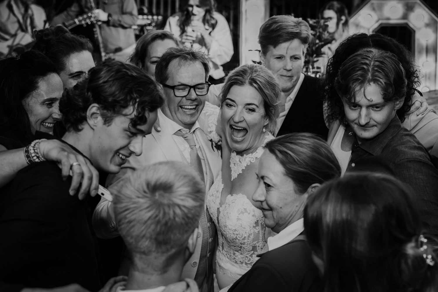 Black and white photo of bride and groom joyfully surrounded by laughing wedding guests in a group hug.