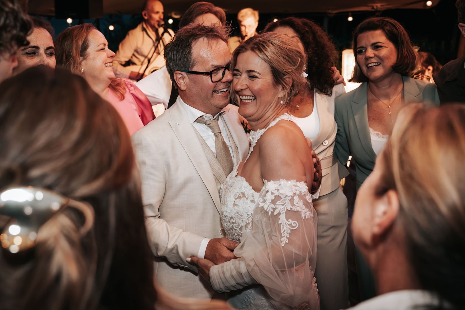 Bride in lace gown and groom in white suit share a joyful moment surrounded by wedding guests.