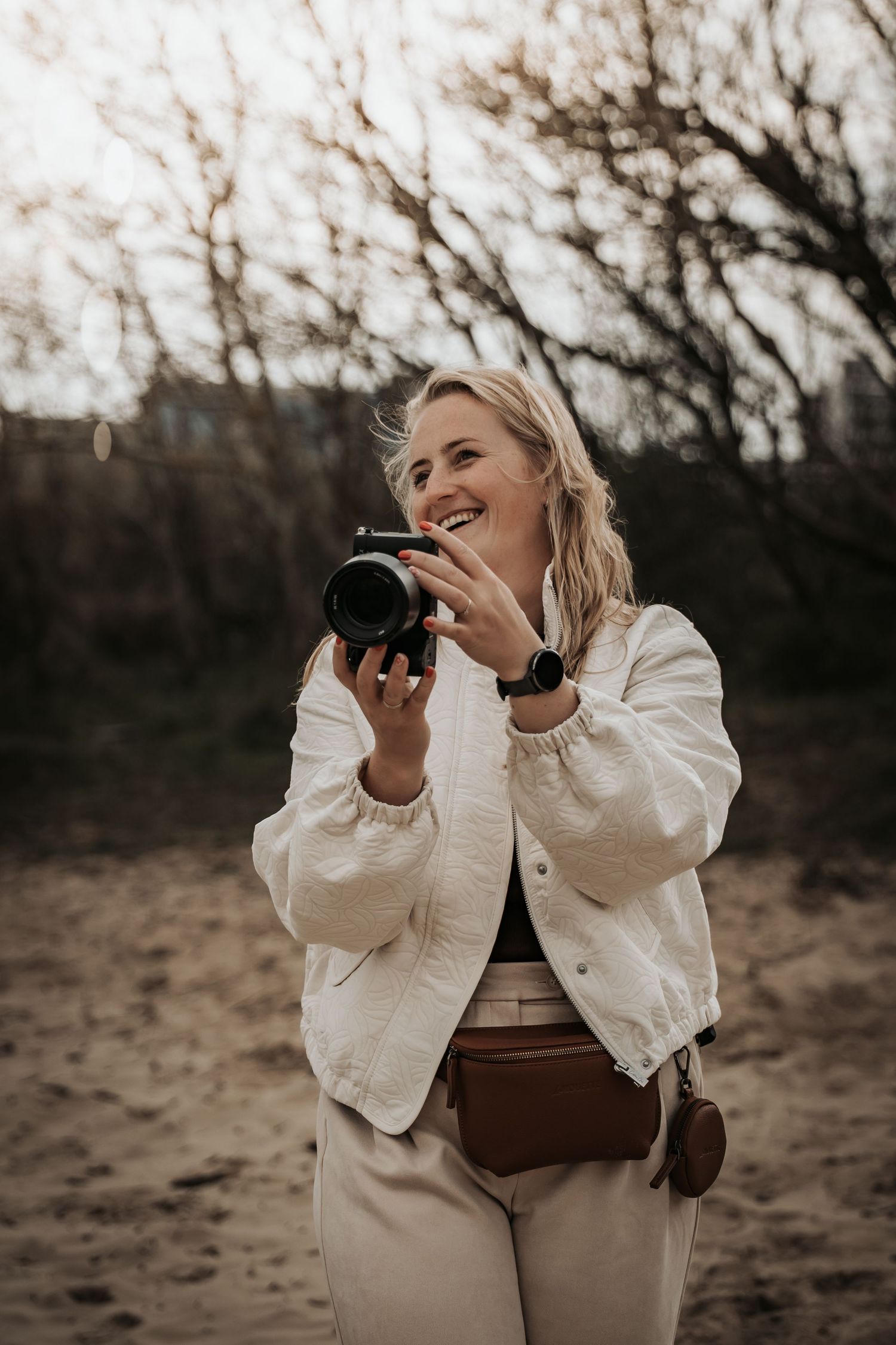 Smiling blonde woman holding a camera outdoors on sandy ground with bare trees in background.