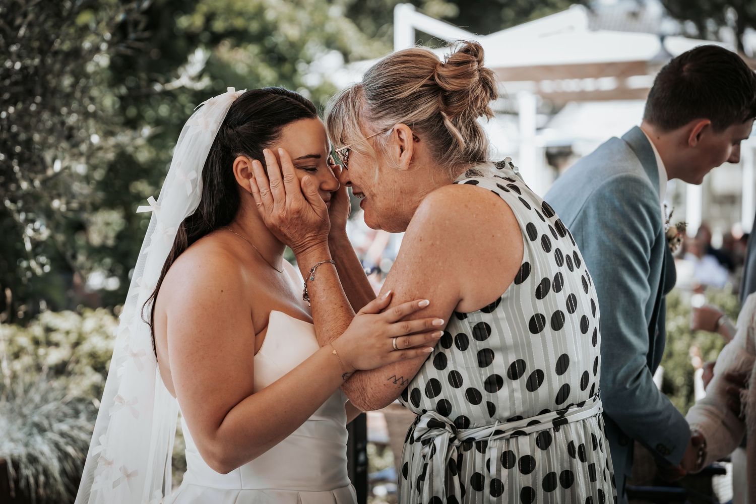 Emotional bride sharing tender moment with older woman, both touching faces at outdoor wedding.