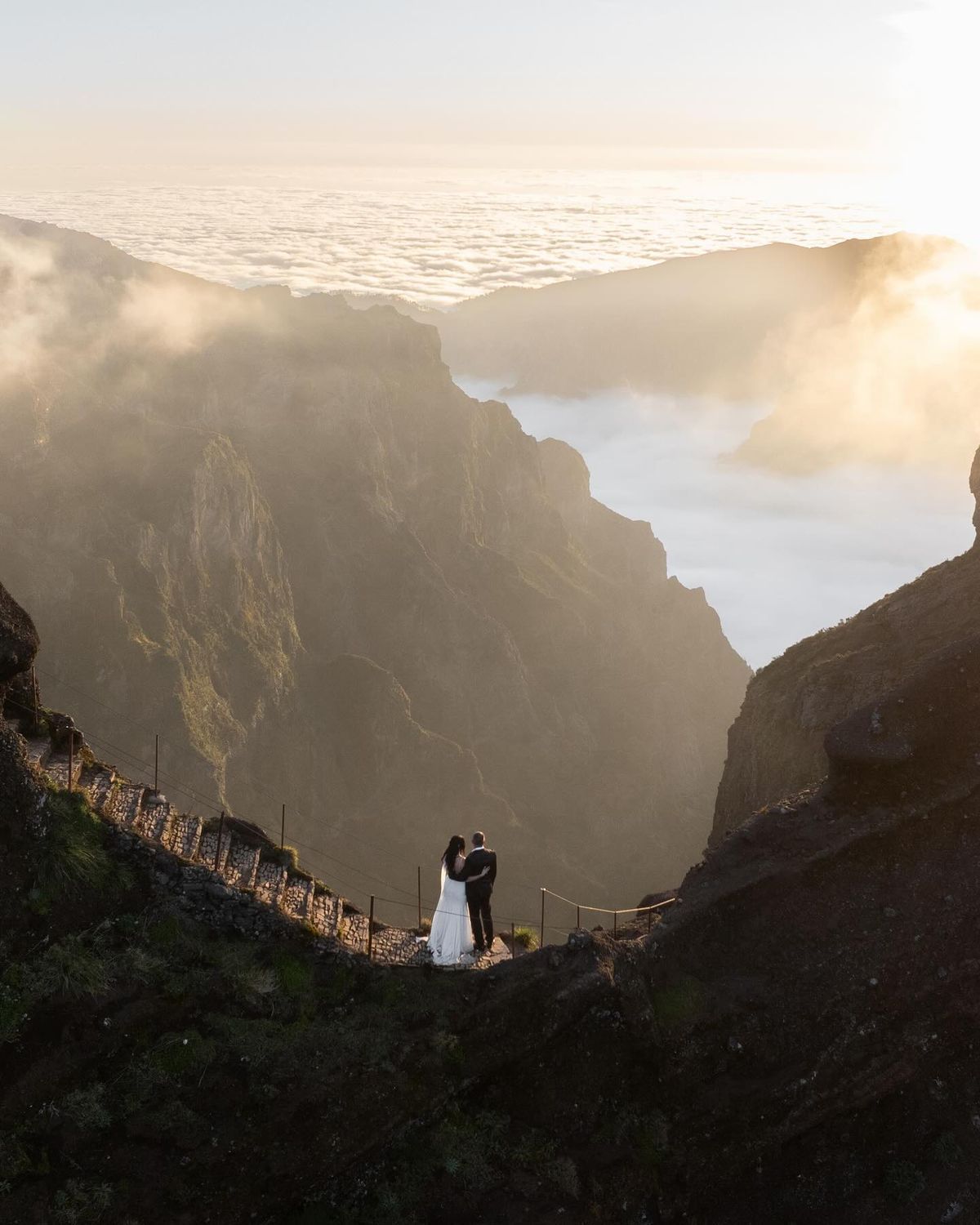 Couple embracing at mountain summit above clouds during golden sunset, dramatic valley and misty peaks in background.