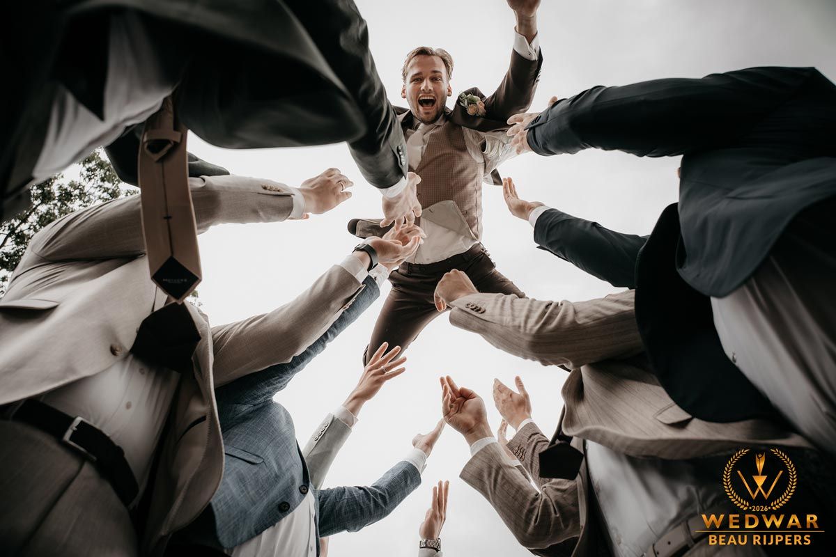 Groomsmen tossing groom into the air in a circle, captured from below at a wedding celebration outdoors.