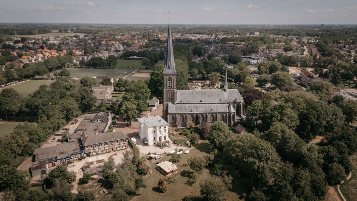 Aerial view of a historic church and white manor building surrounded by trees in a town.