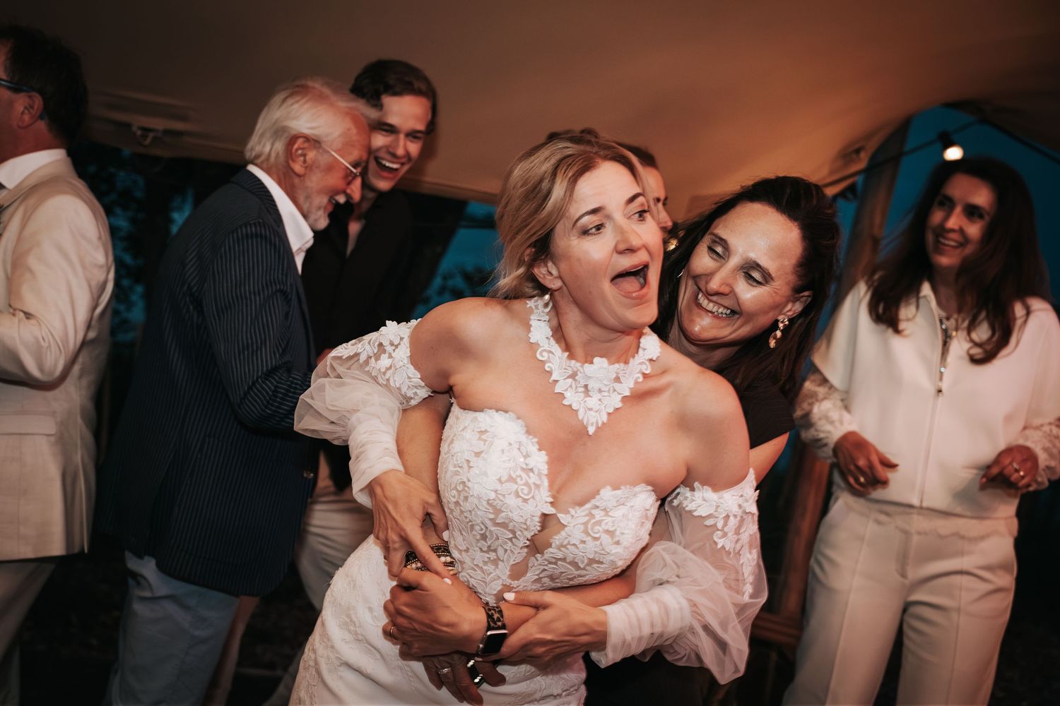 Bride laughing joyfully while being hugged by a guest at a lively wedding reception party.