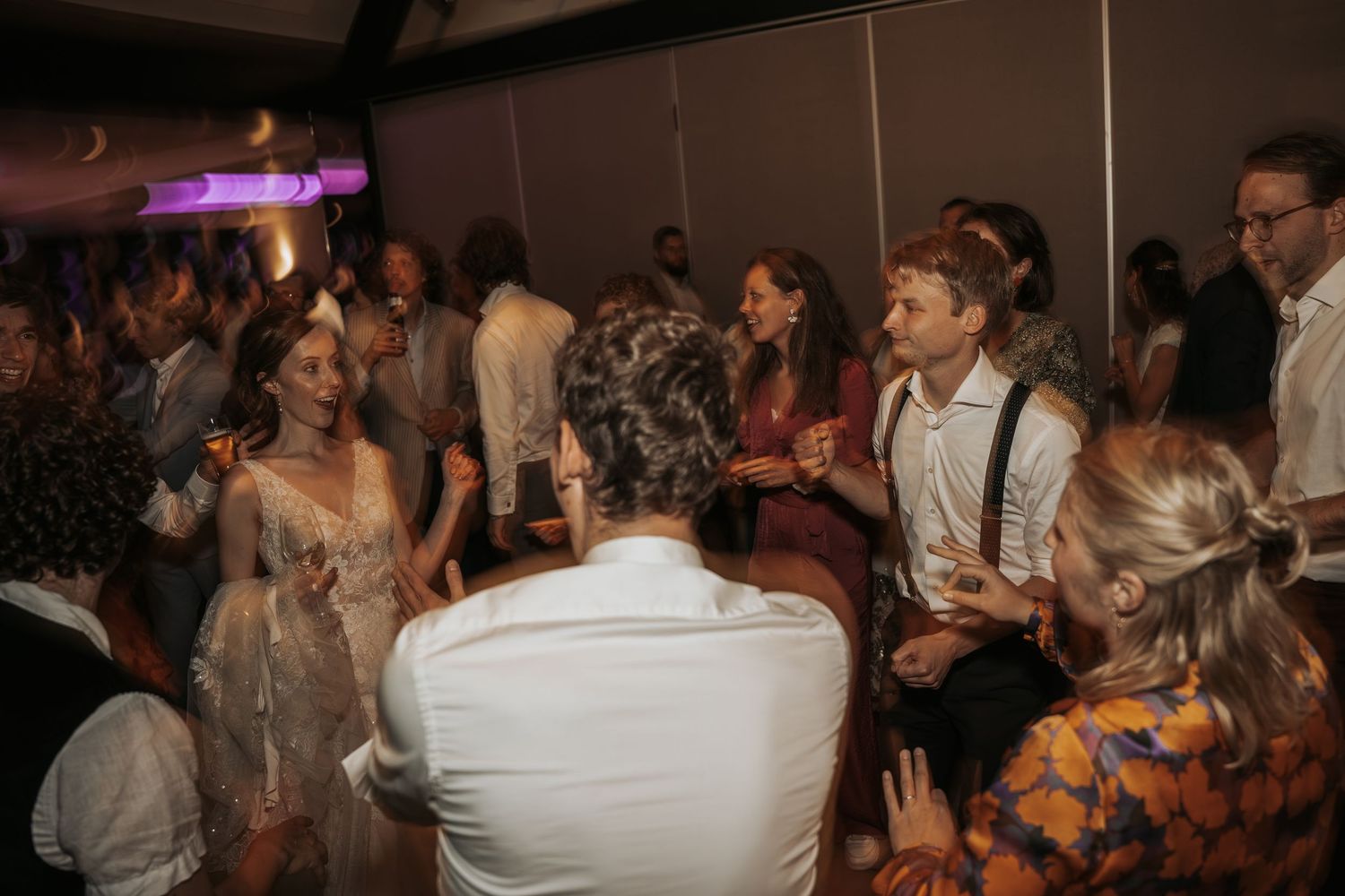 Wedding guests mingling and dancing together in a warmly lit reception venue with purple lighting.
