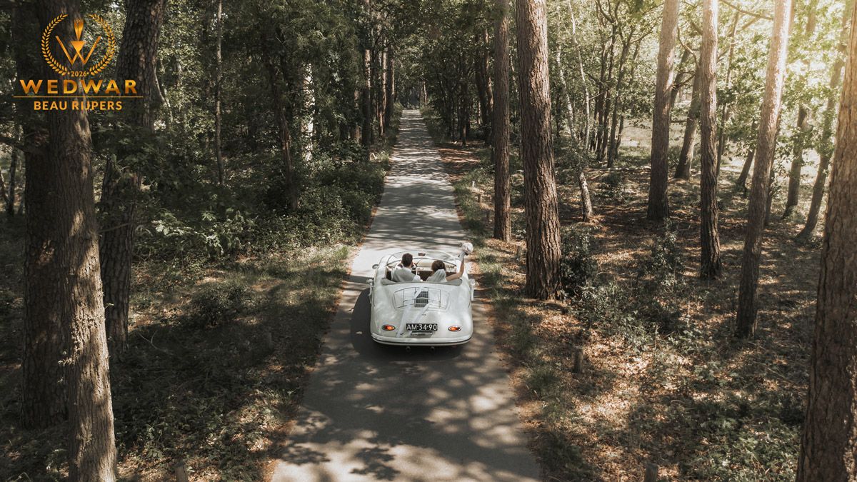 A vintage white convertible car drives down a tree-lined forest path, captured from an aerial drone perspective.
