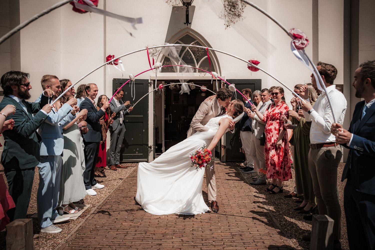 Bride and groom sharing dramatic dip kiss under ribbon arch with cheering wedding guests.