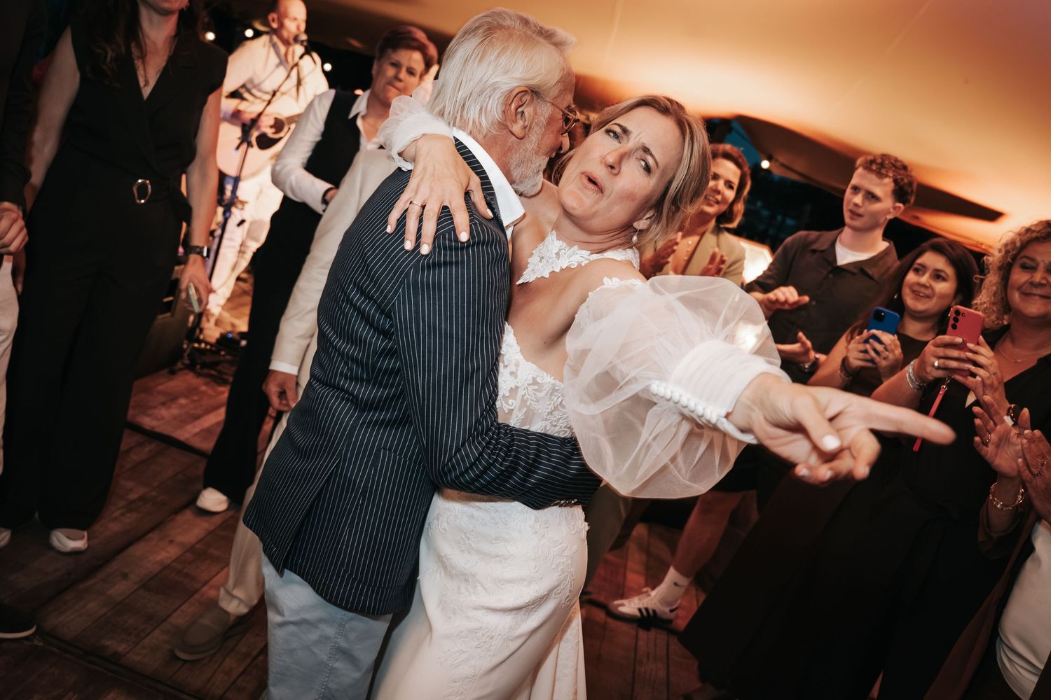 Bride in white gown dances joyfully with an older gentleman at a warmly lit wedding reception.