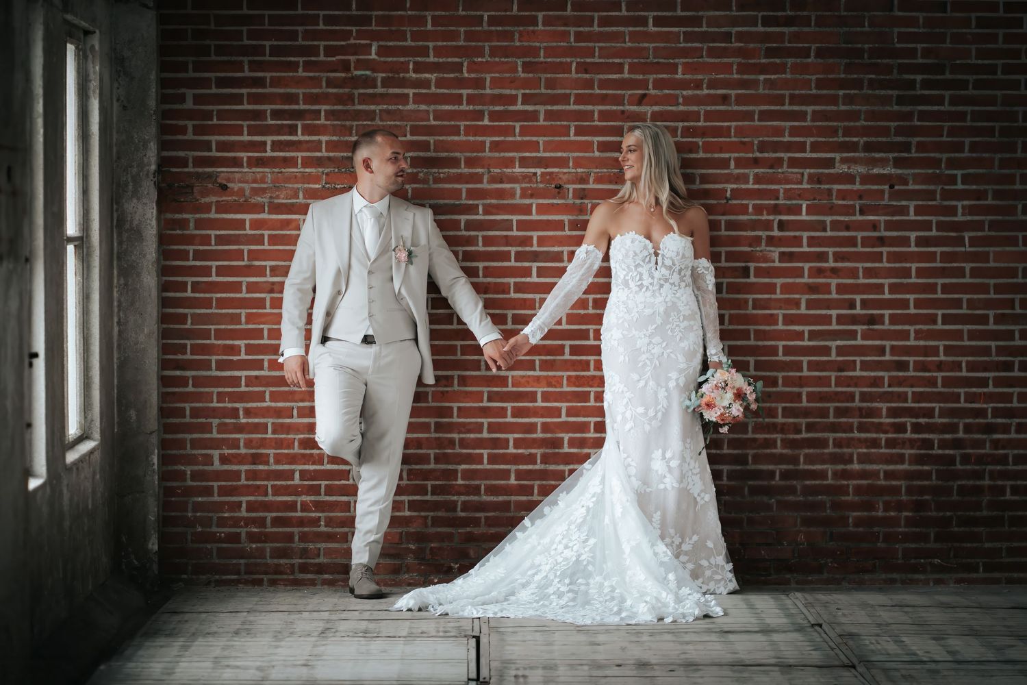 Bride and groom holding hands in front of a red brick wall, wearing elegant white wedding attire.