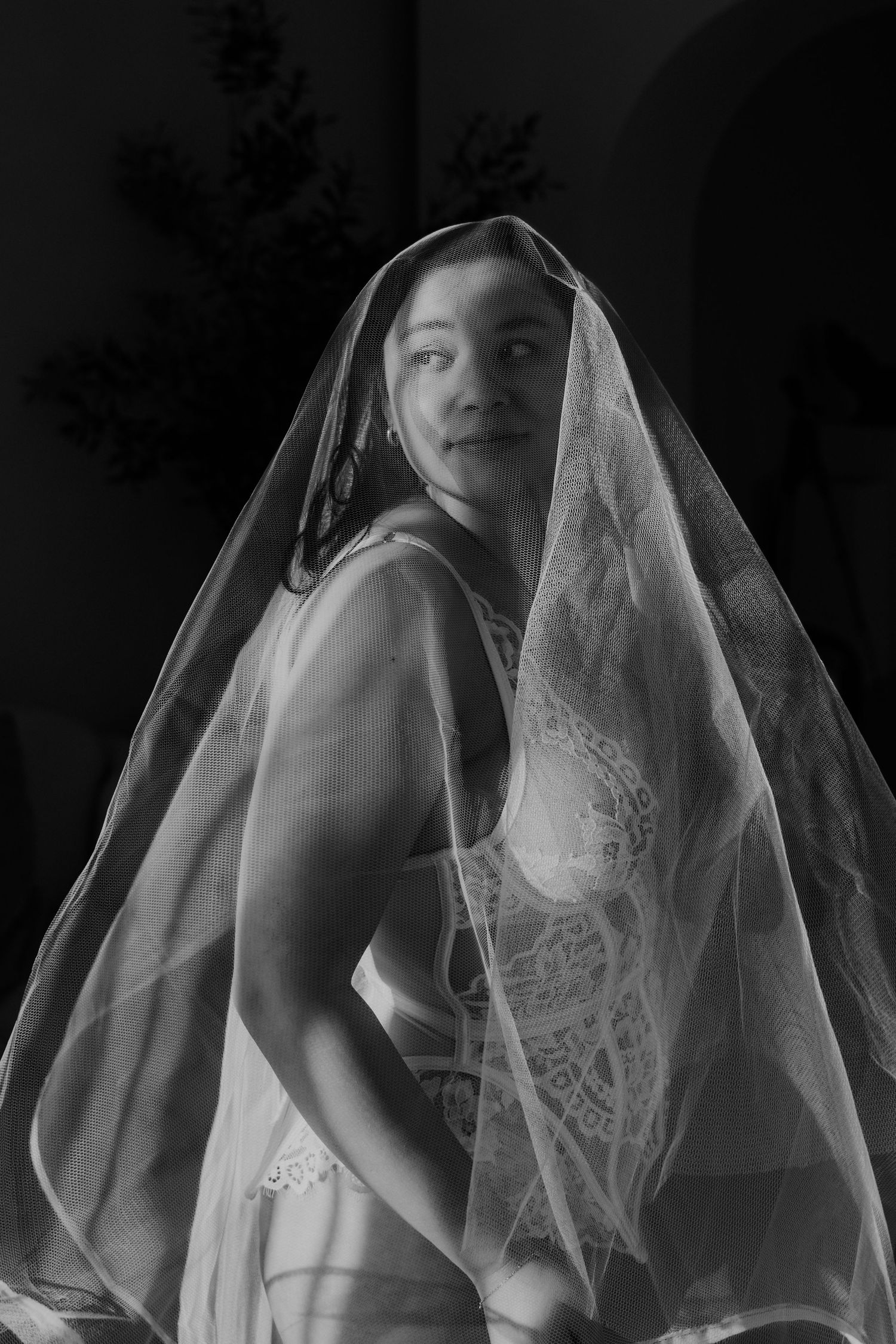 Black and white portrait of woman wearing delicate lace veil, smiling softly in dim light.