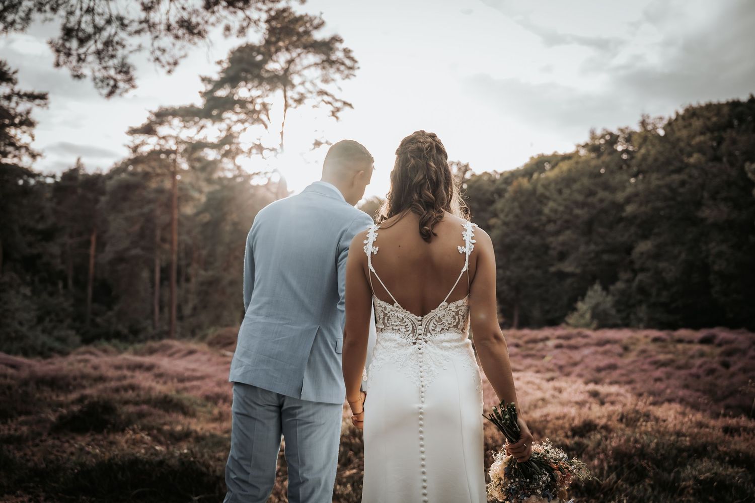 Bride in lace-back gown and groom in light blue suit walking through blooming heather forest.