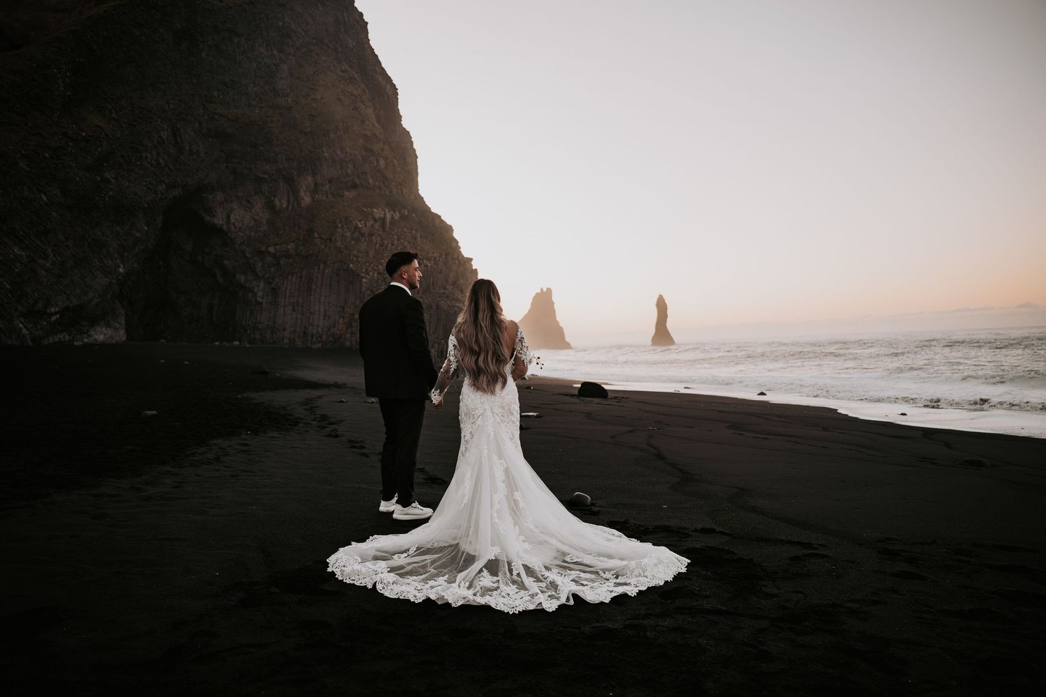Couple in wedding attire stand on black sand beach at sunset with dramatic sea stacks in background.