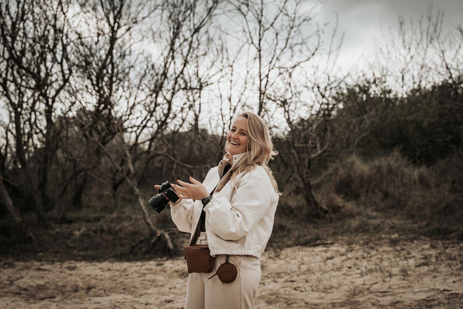 Smiling blonde woman holding a camera outdoors in a natural winter landscape.