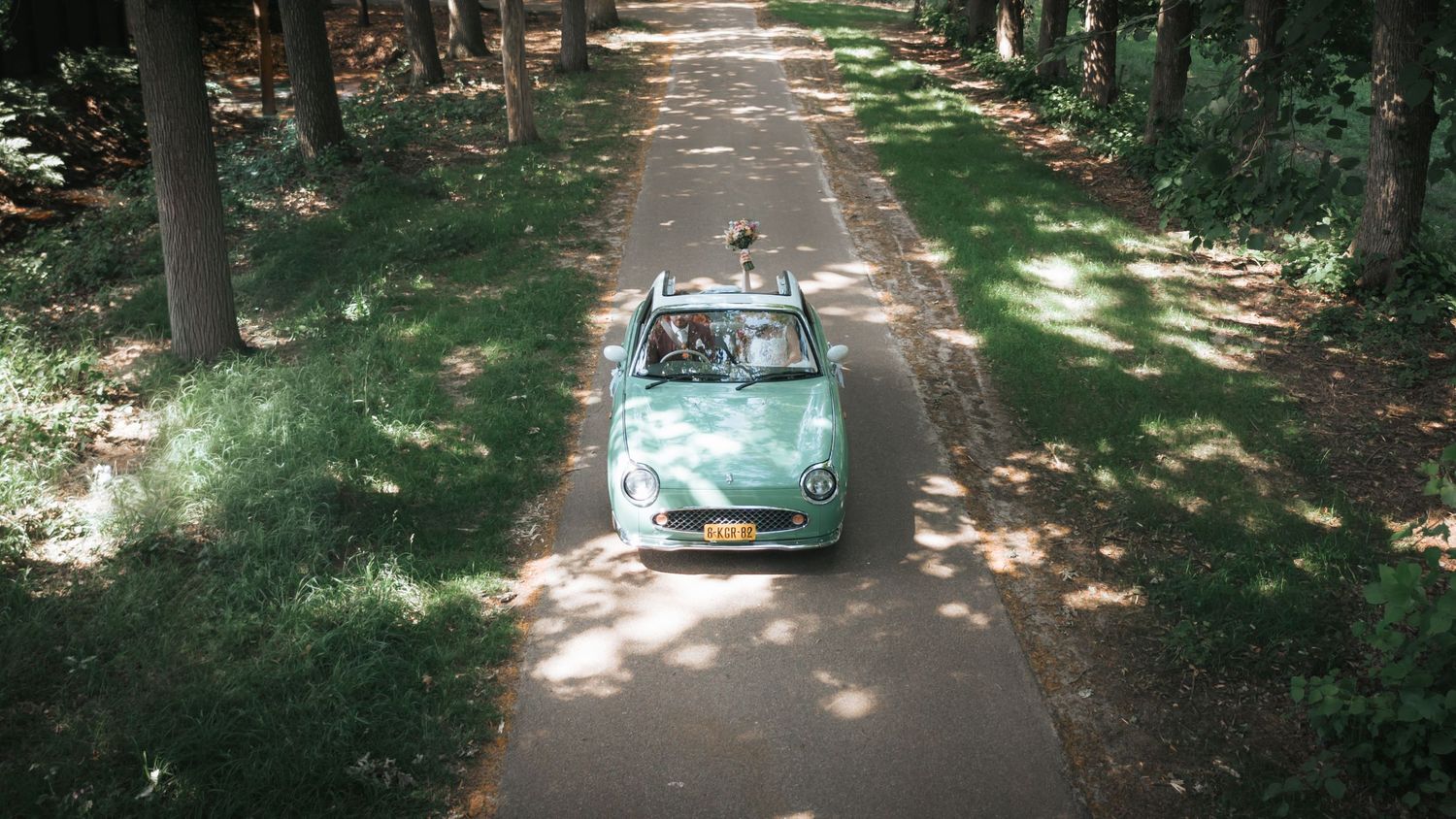 Aerial view of a classic mint green convertible driving down a sunlit tree-lined wedding day road.