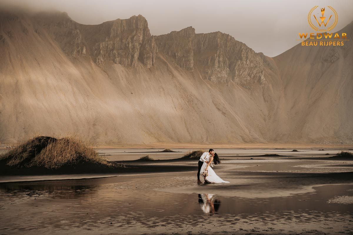 Couple embracing on black sand beach with dramatic misty mountains backdrop during wedding photoshoot in Iceland.