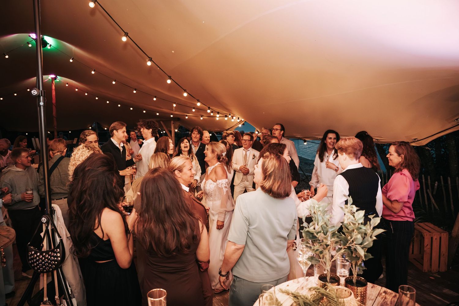 Guests gathering and dancing together inside a warmly lit tented wedding reception venue.