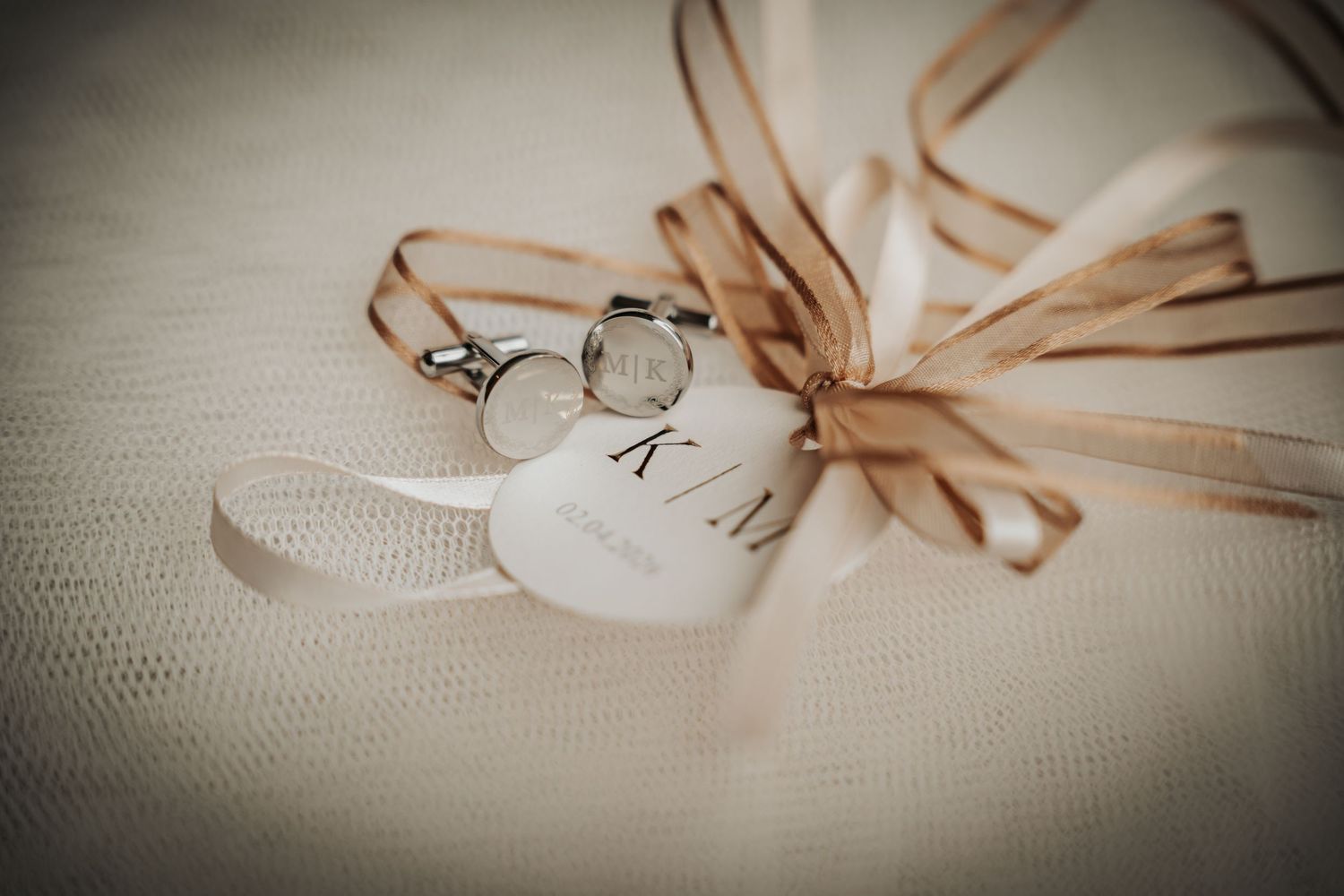 Wedding rings and a heart-shaped tag with initials tied with elegant ribbon on white fabric.