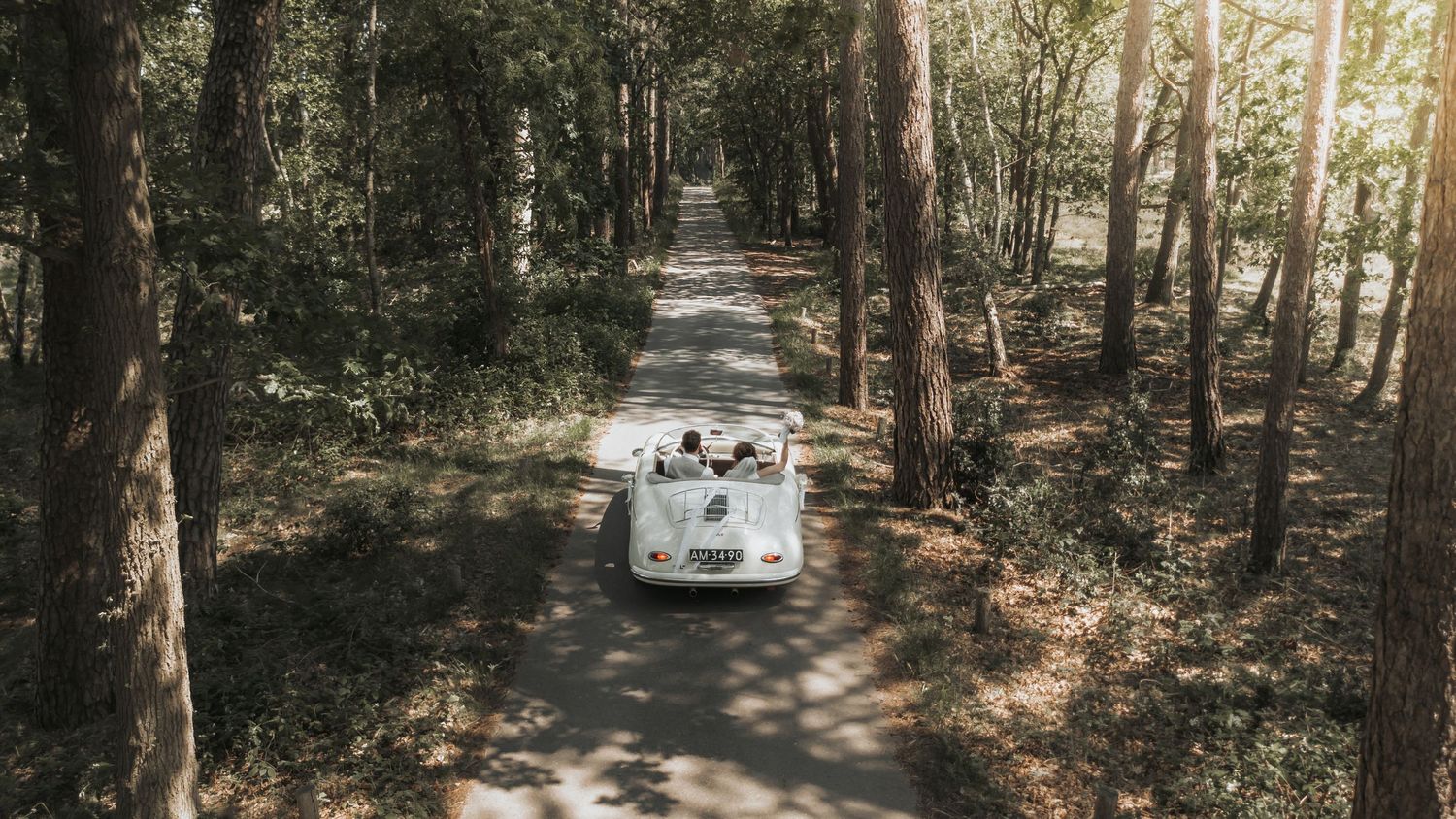 Vintage white convertible car driving through a sunlit forest path on a wedding day.