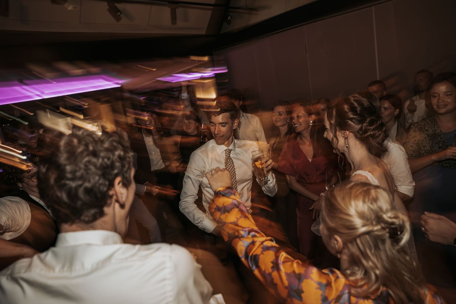 Energetic wedding dance floor scene with guests celebrating under purple lights at a reception.