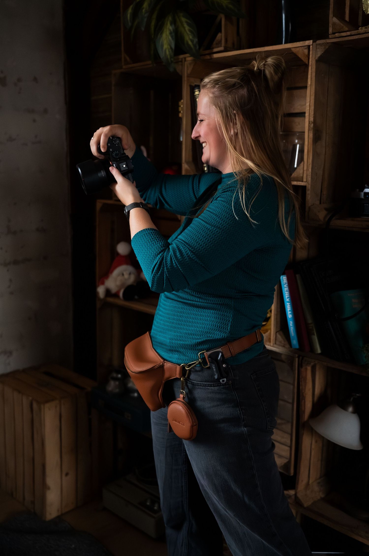 Smiling blonde woman in teal top holding DSLR camera, wearing leather belt bag, standing near rustic wooden shelving unit.
