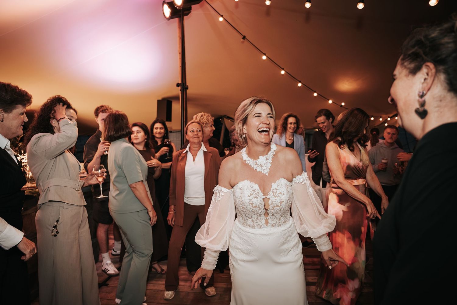 Bride in a white off-shoulder gown dancing and laughing at an elegant tented wedding reception.