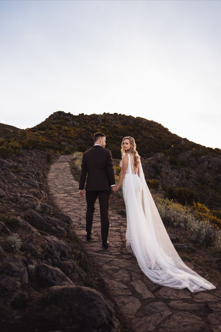 Bride in white gown with long train holds groom's hand on stone path with scenic hills backdrop.