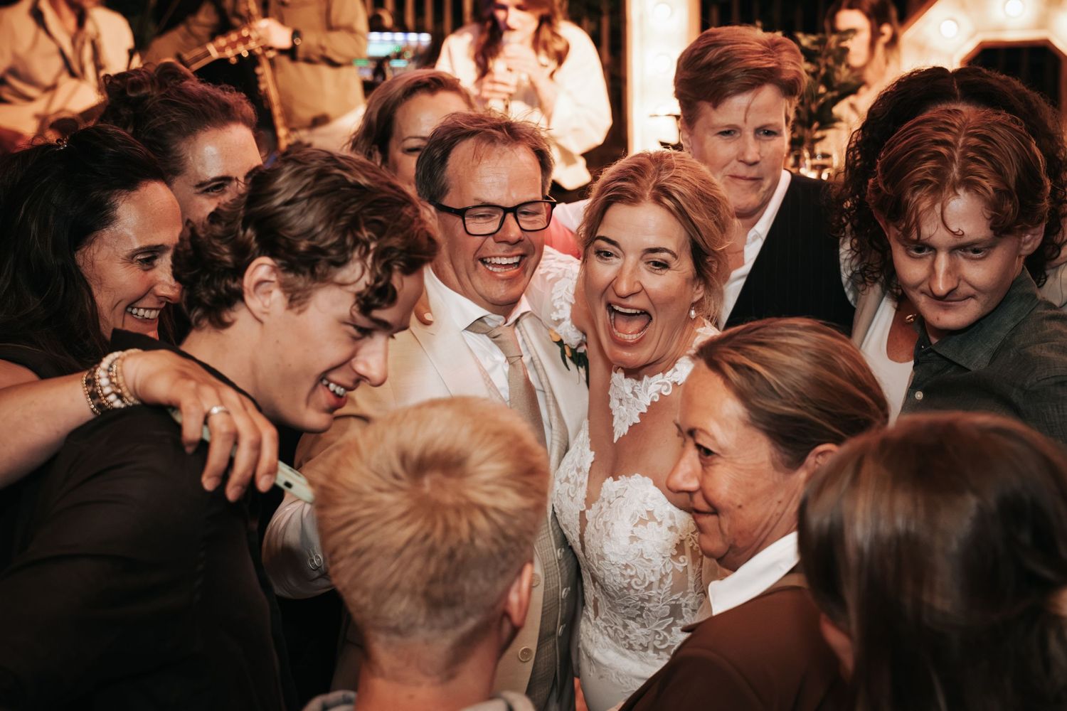 Bride and groom beaming with joy as wedding guests gather around them in a warm group embrace.
