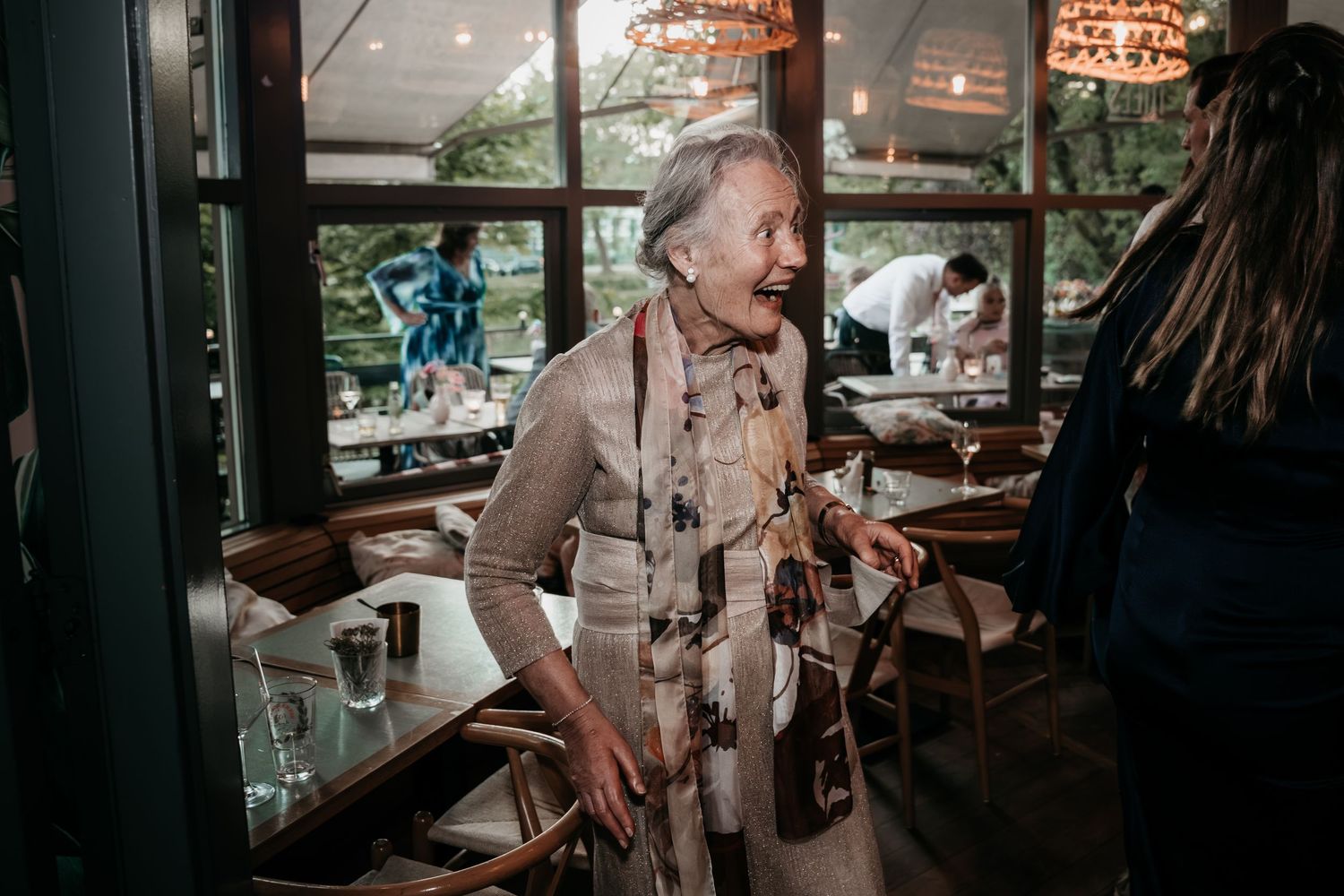 Elderly woman smiling warmly while holding a bottle at an elegant indoor wedding reception venue.