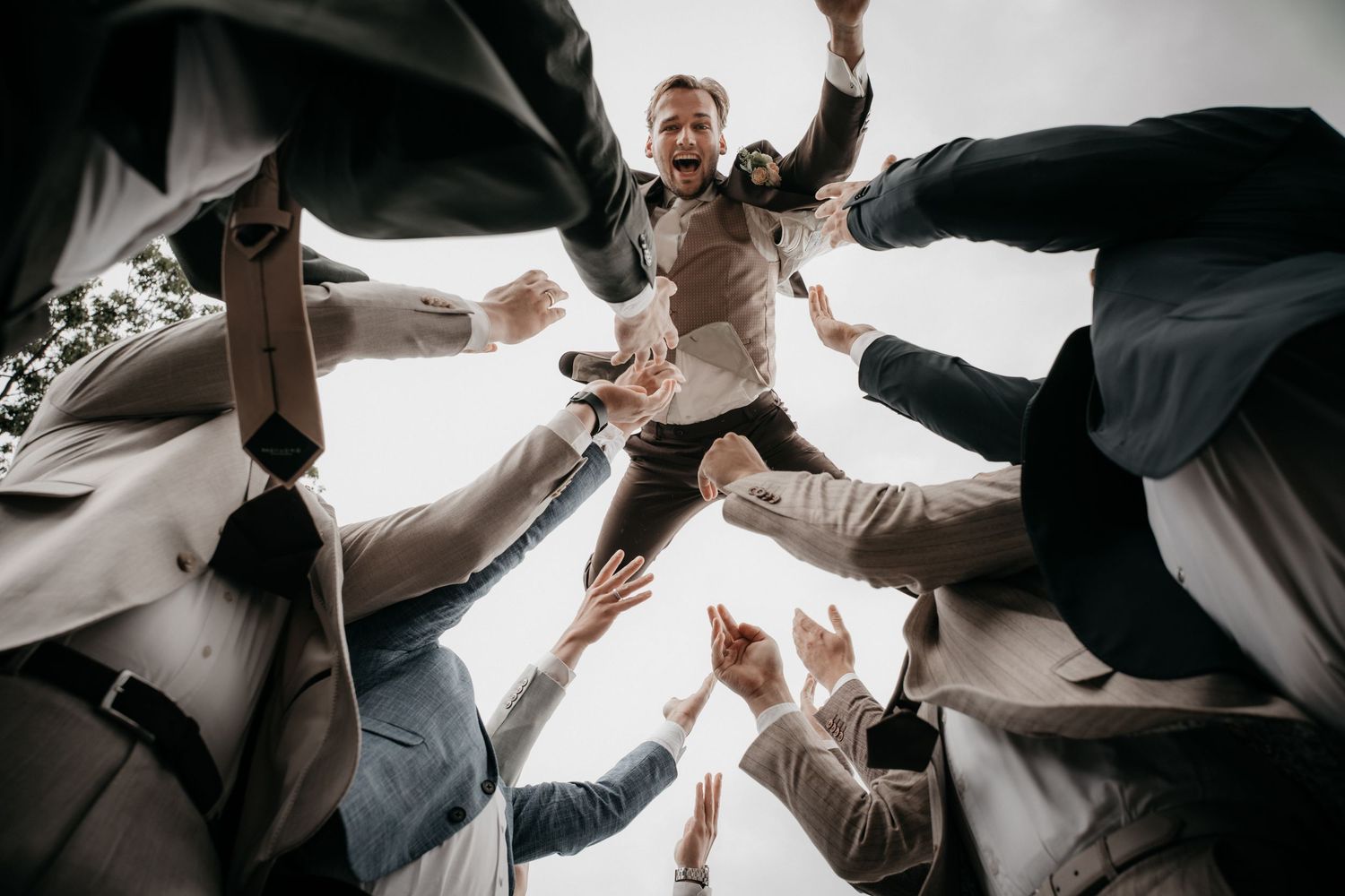 Groomsmen tossing the groom into the air in a joyful wedding celebration outdoors.