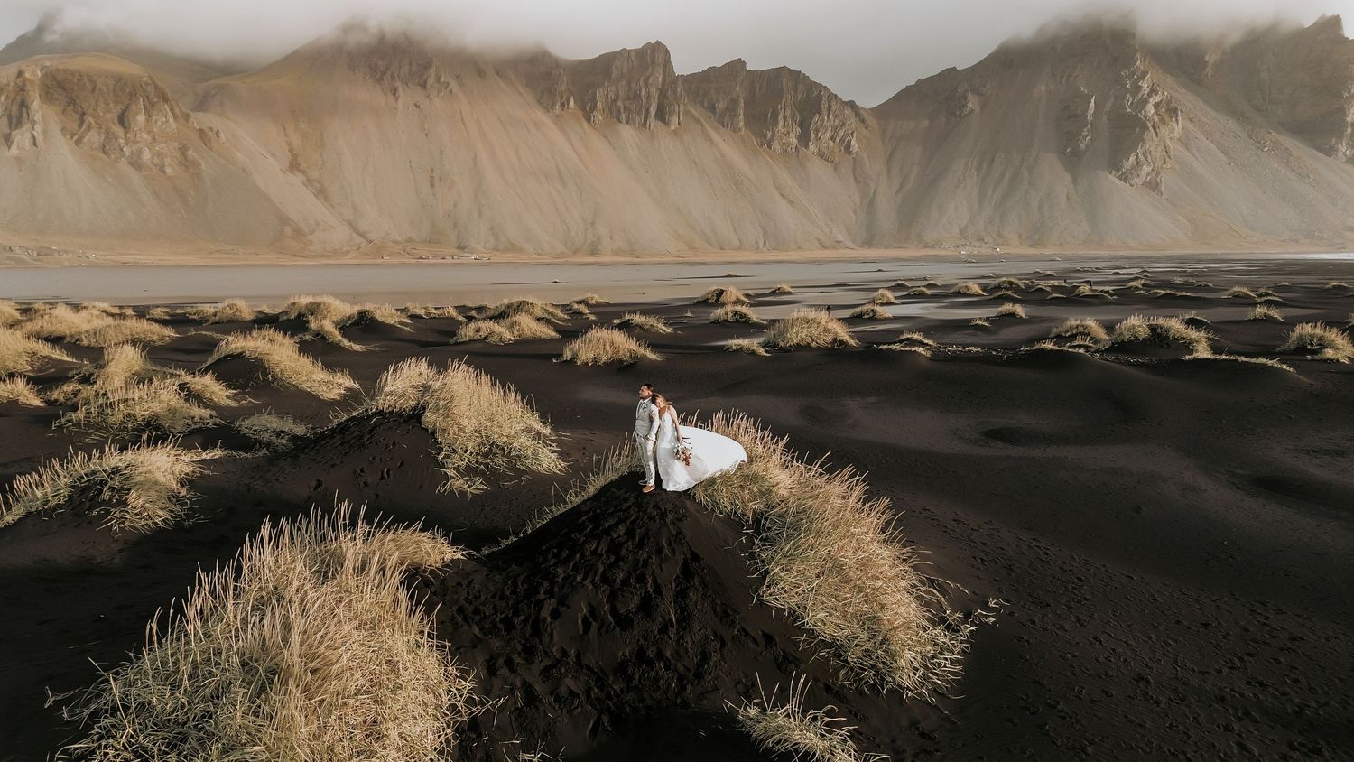Aerial view of a bride and groom standing on dramatic black sand dunes with misty mountains.