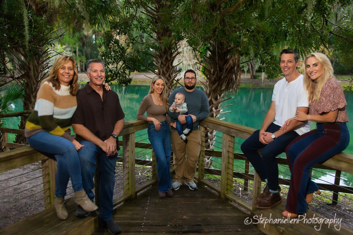 Family of six posing on a wooden bridge surrounded by lush green trees and a turquoise lake during a professional photo shoot.