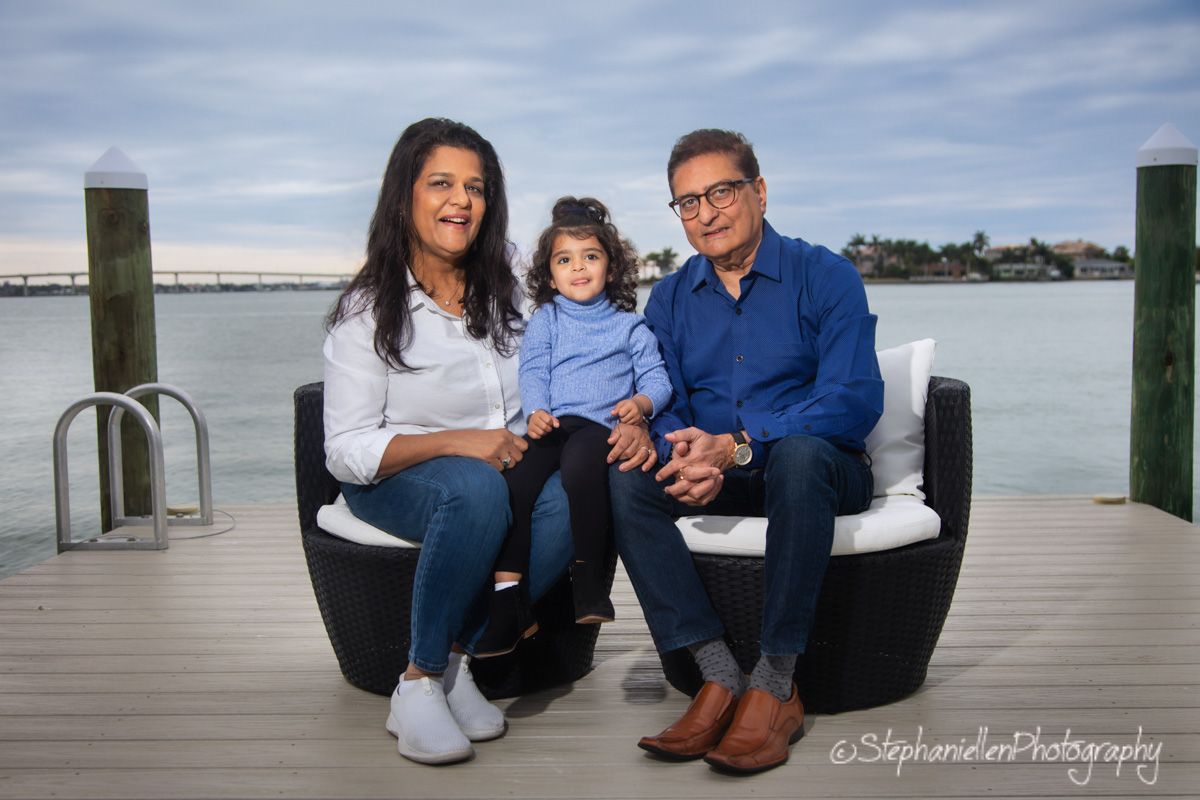 Family of three sitting on outdoor furniture on a waterfront dock, parents with young child.
