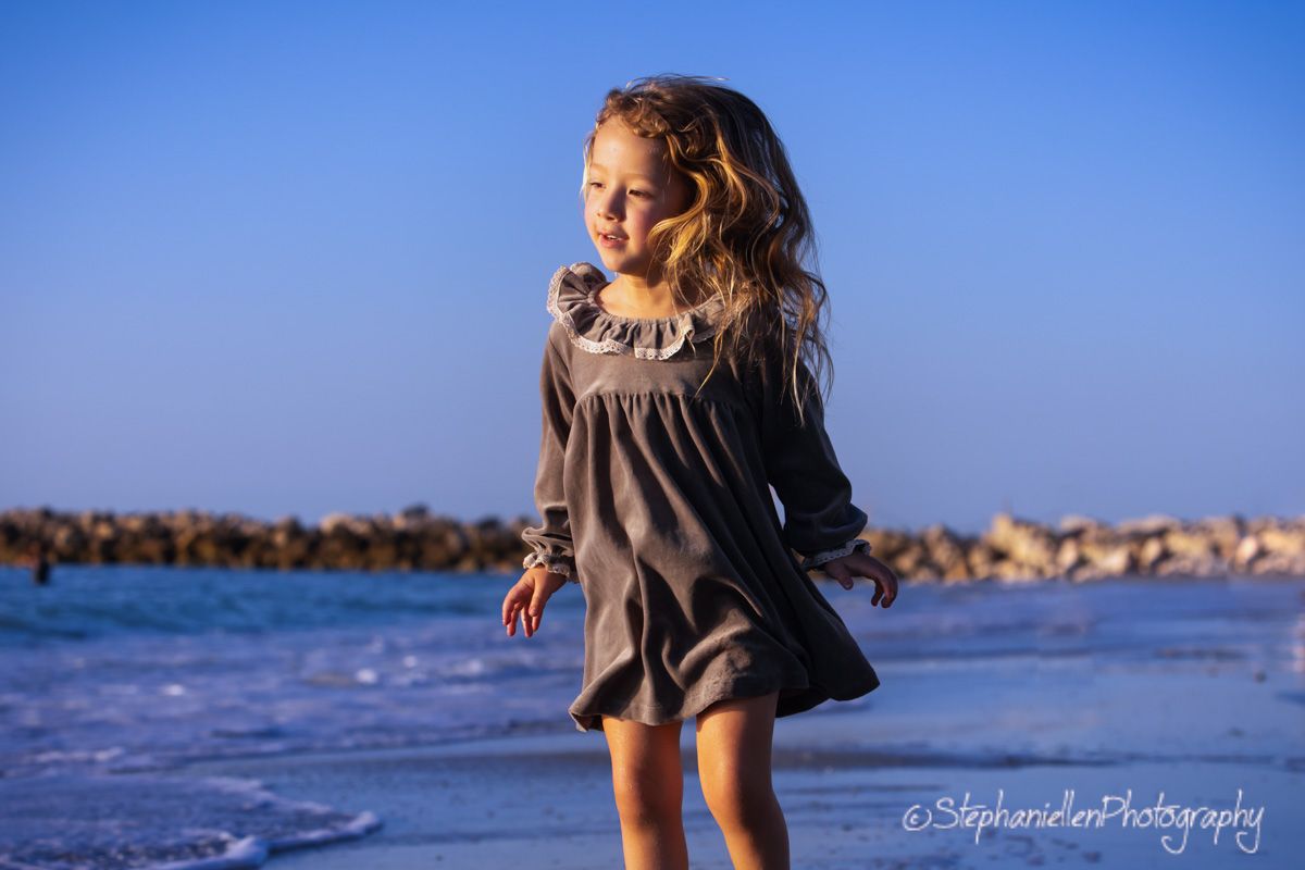 Young girl in gray dress twirling on a beach with blue sky and ocean waves in the background.