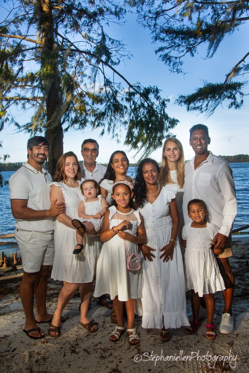 Large extended family in matching white outfits posing together on a sunny lakeside beach.