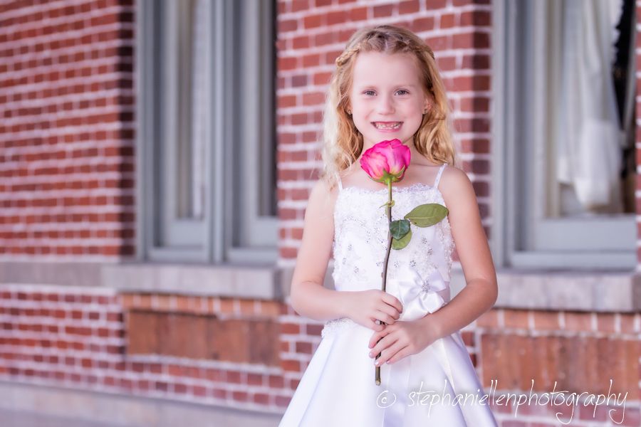 Young girl in white dress holding a pink rose, smiling outdoors near a brick building.
