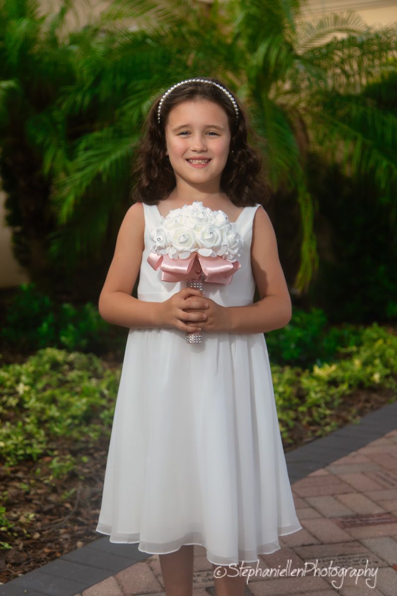 Young flower girl in a white dress holding a white floral bouquet with a pink ribbon handle.