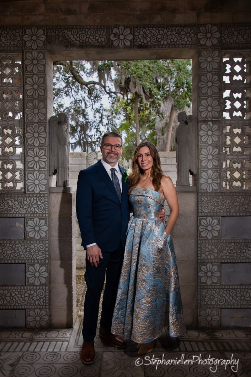 Bride in blue floral gown and groom in navy suit pose in ornate stone doorway.