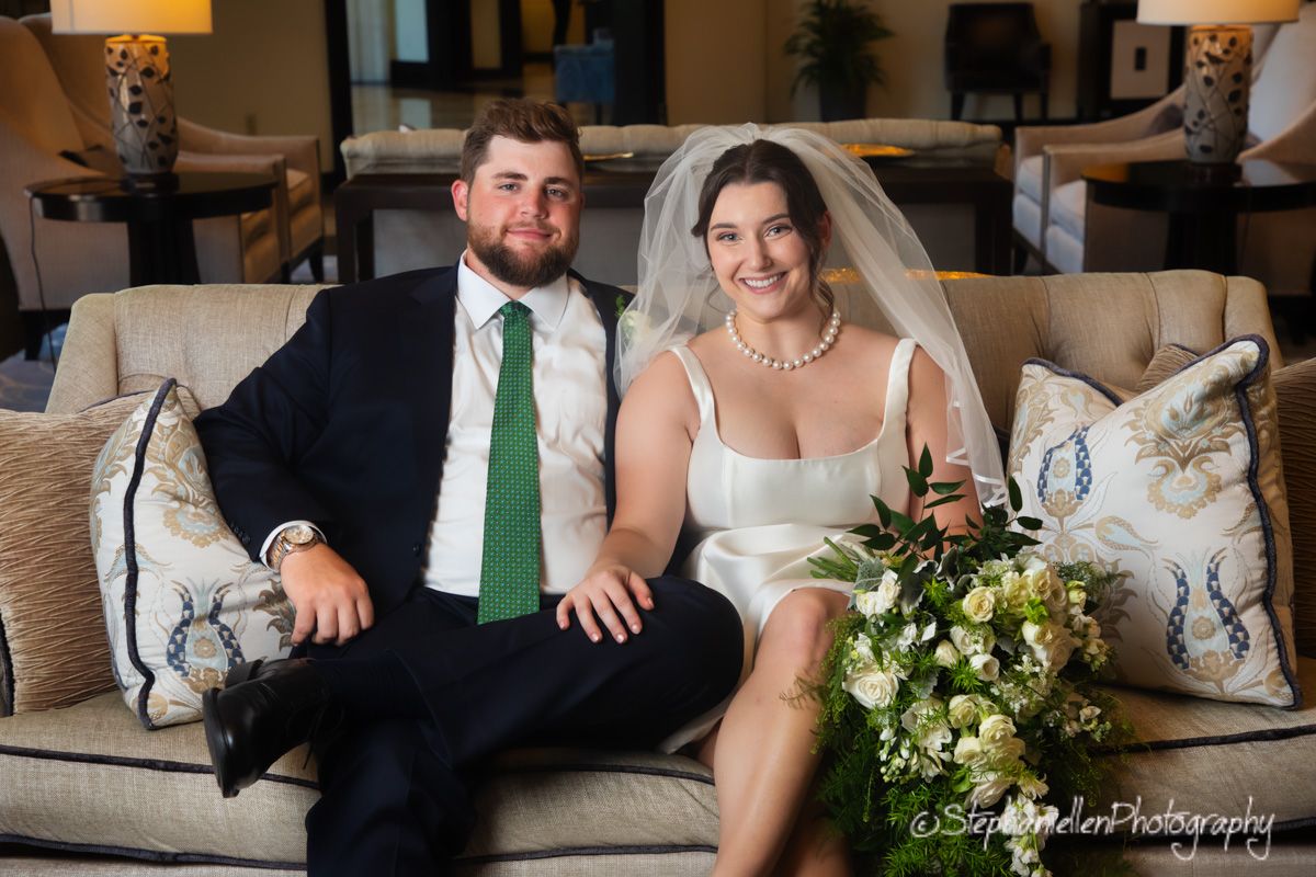 Bride and groom sitting together on an elegant sofa, bride holding a white floral bouquet.