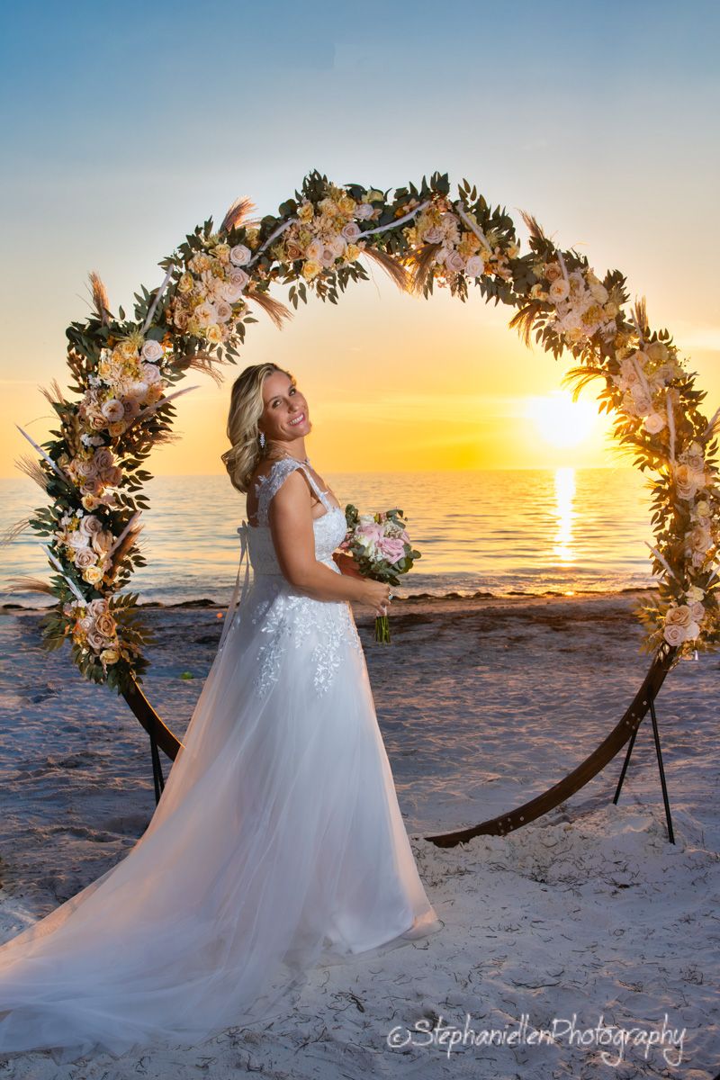 Bride in white gown stands under floral arch at beach sunset wedding.