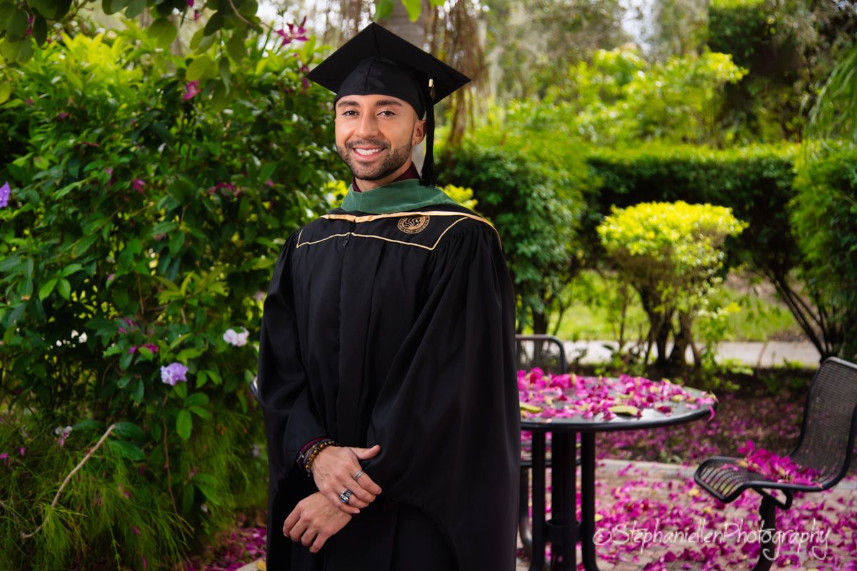 Graduate in black cap and gown smiling in colorful garden setting.