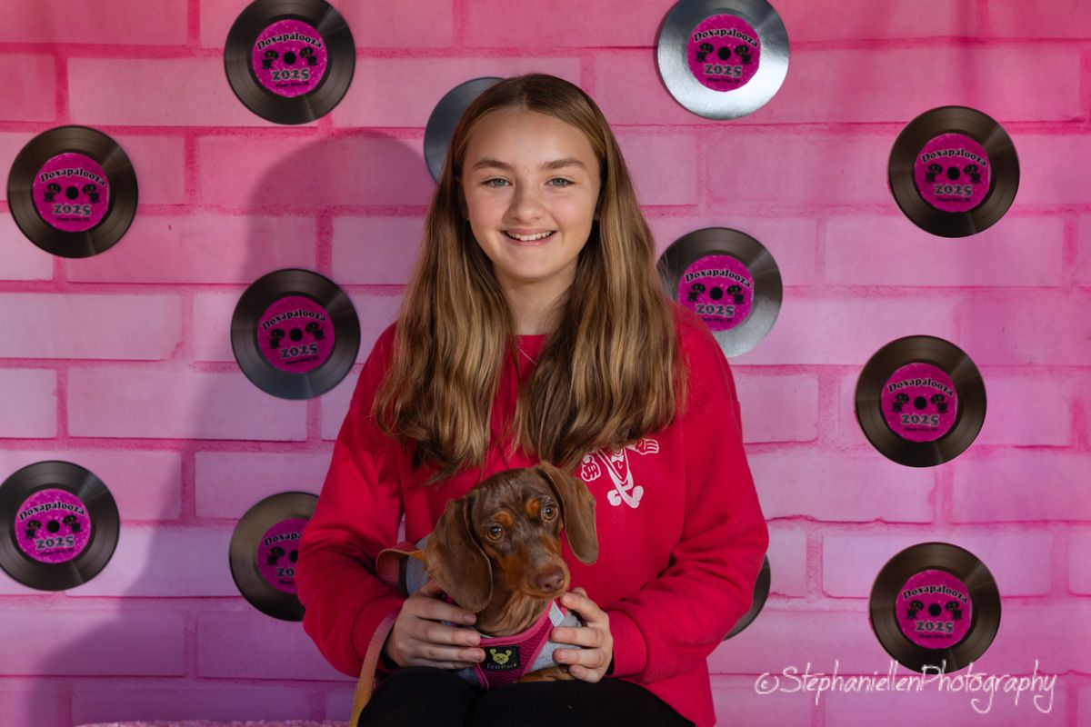 A smiling teenage girl in a red hoodie holds a small dachshund puppy in front of a pink wall with vinyl record decorations.