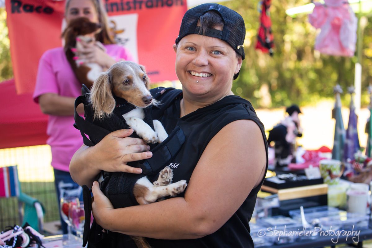 Woman smiling and holding a small dog dressed in costume at an outdoor charity event with pink decorations.