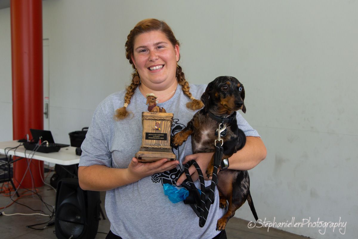 Woman smiling while holding a dachshund dog and a trophy award at an indoor event.